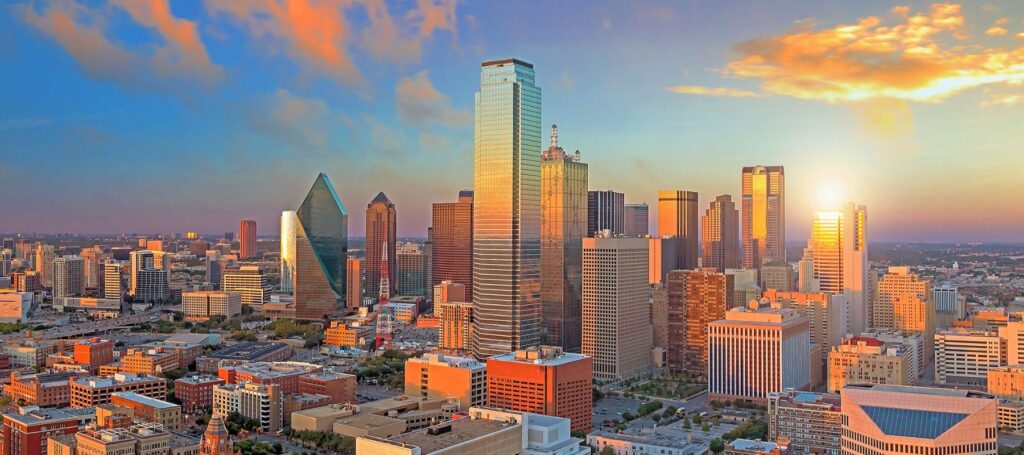 n aerial view of the Dallas skyline at sunset shows a cluster of modern skyscrapers reflecting the orange and blue sky