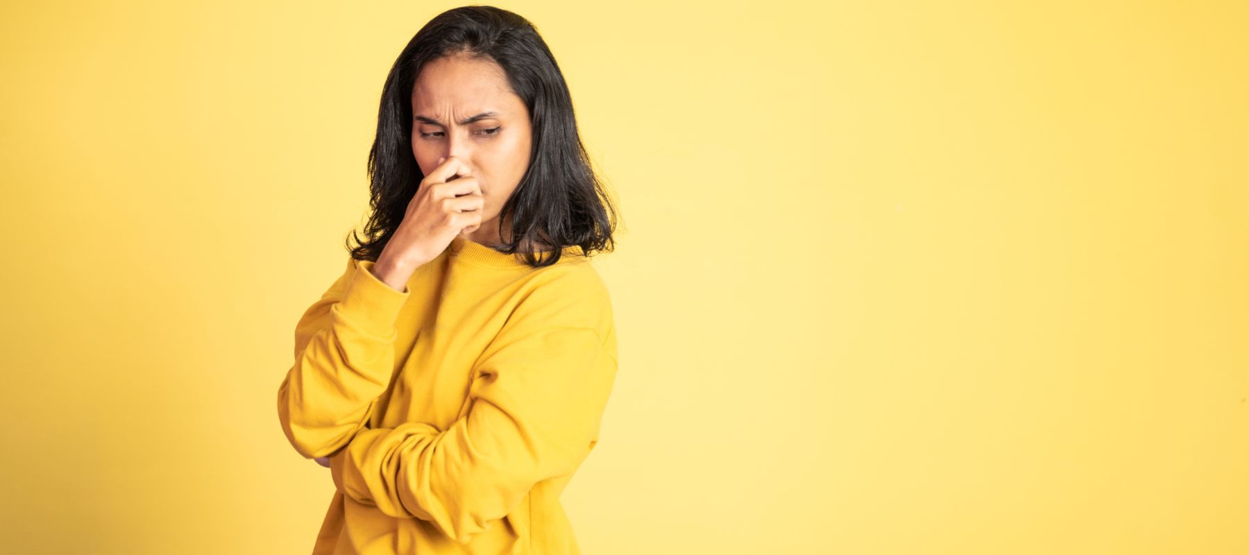 A woman in a yellow sweater pinches her nose with a disgusted expression on a yellow background