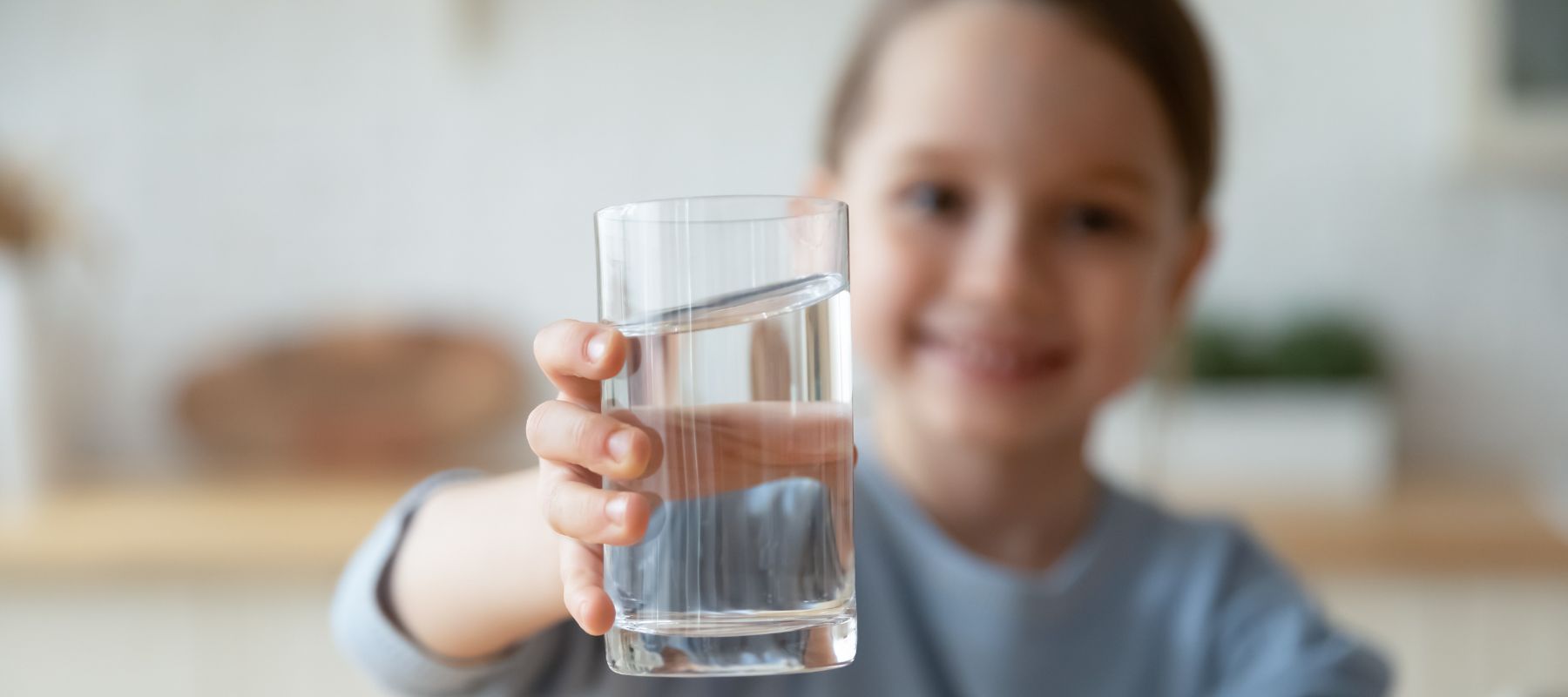 A young girl holds a glass of water out to the camera, with a smile blurred in the background