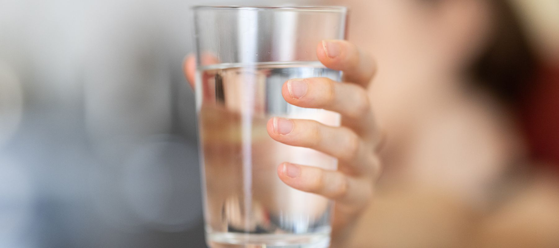 A person holds a glass filled with clear water, with a blurred background