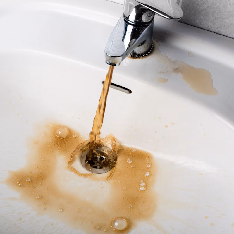 Rusty, brown water pours from a faucet into a white sink, staining the basin