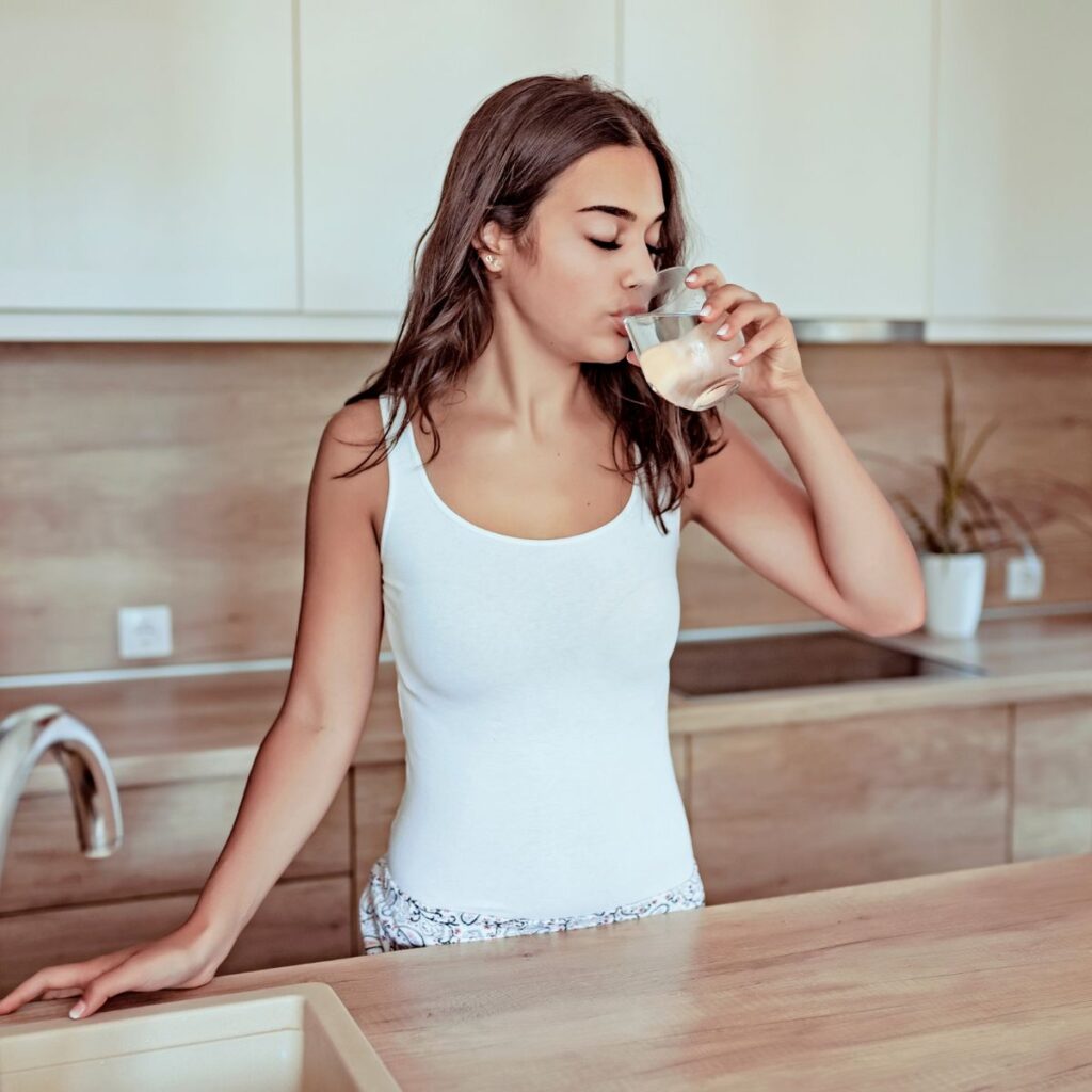 A woman in a white tank top sips from a glass of water while leaning against a wooden kitchen counter