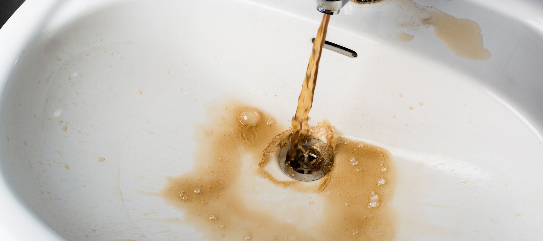 Rusty, brown water pours from a faucet into a white sink, staining the basin