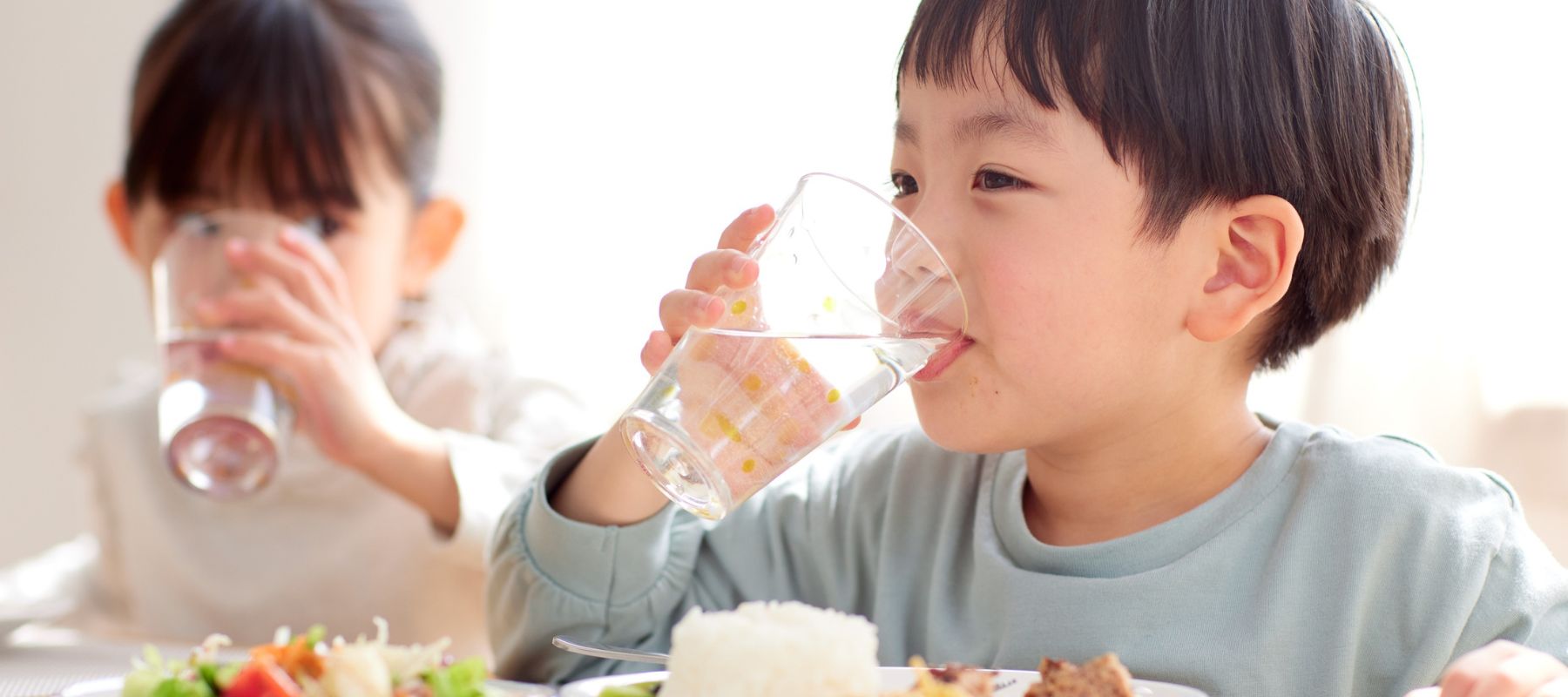 A young boy is drinking from a glass of water while sitting at a table with a meal in front of him