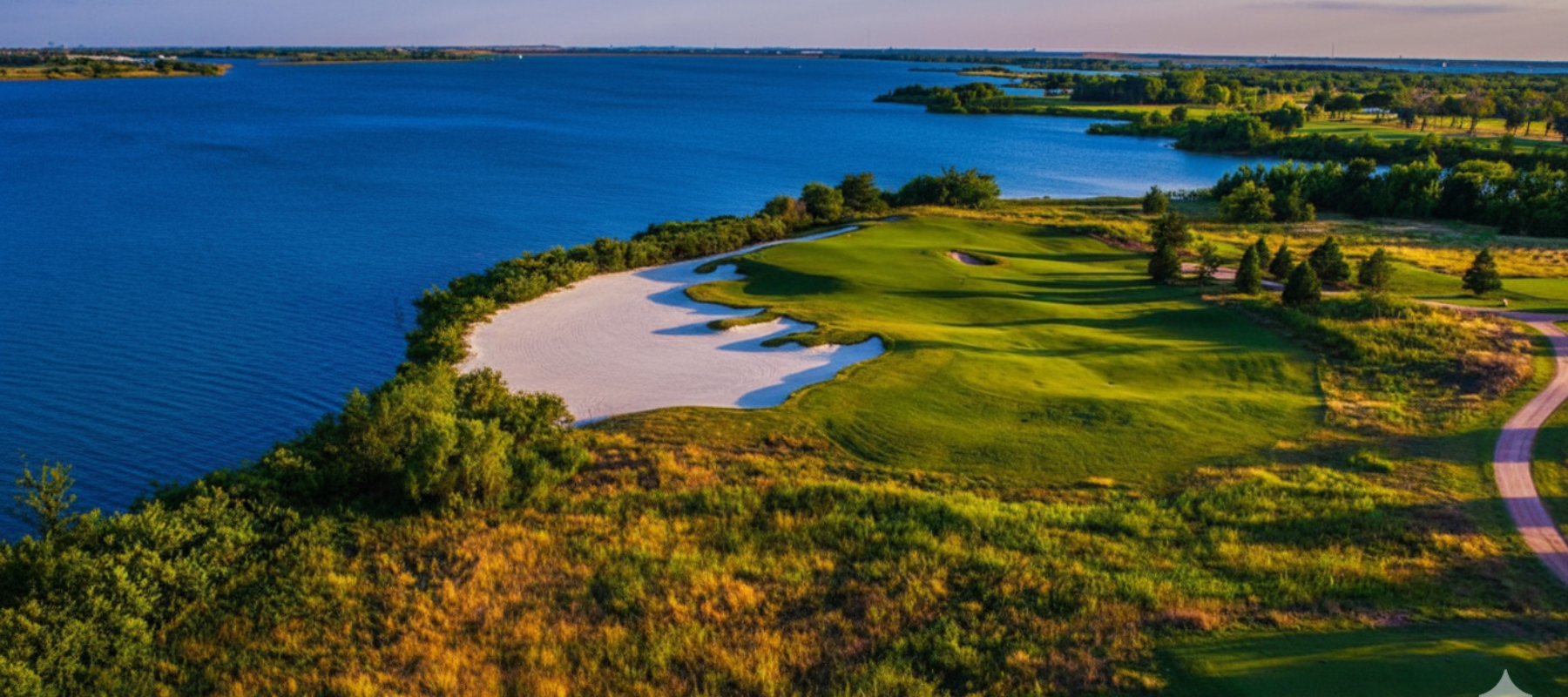 A scenic lakeside golf course in The Colony, Texas, featuring a white sand bunker and rolling green fairways along the blue water at sunset.