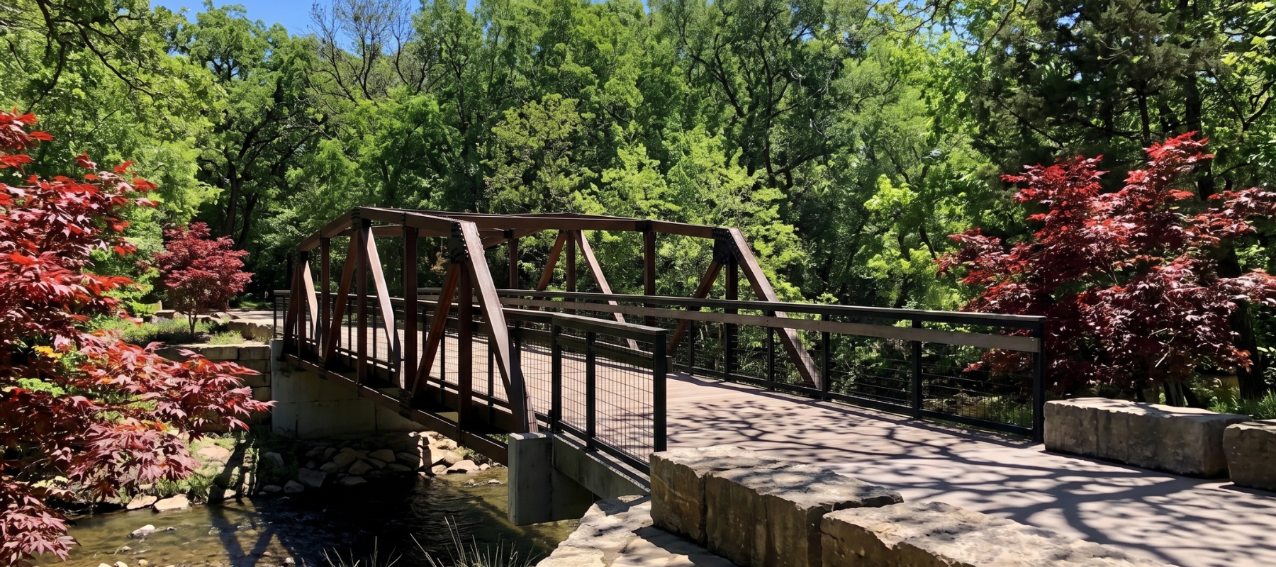 A brown metal truss pedestrian bridge spans a quiet creek in a lush, sunlit park. The bridge is framed by vibrant red Japanese maples in the foreground and a dense canopy of green trees in the background. Large stone blocks line the paved walkway leading onto the bridge. At Prairie Creek Park in Richardson, Texas.